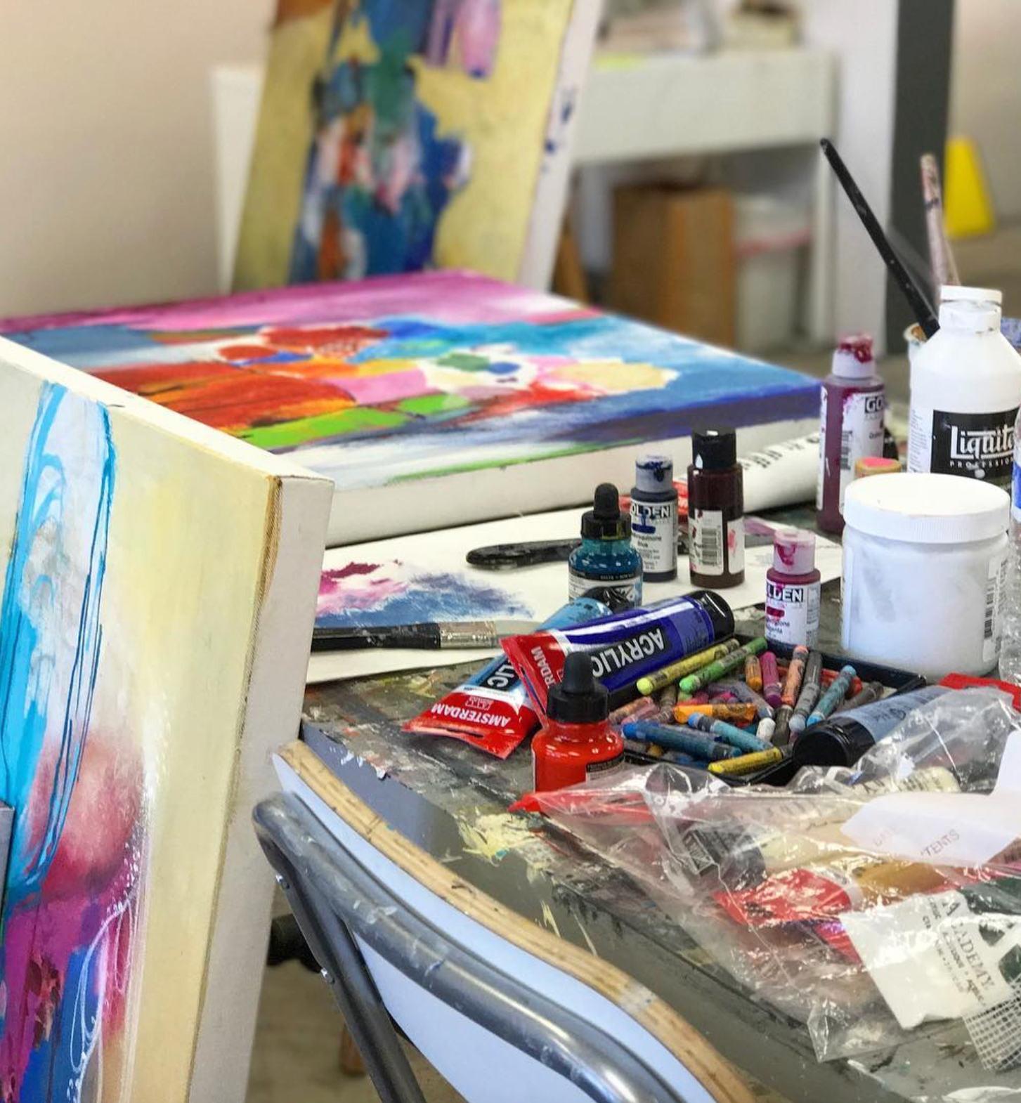 A cluttered artist's studio table with multiple colorful paintings on easels, paint tubes, brushes, water bottles, and other art supplies in a real indoor environment.